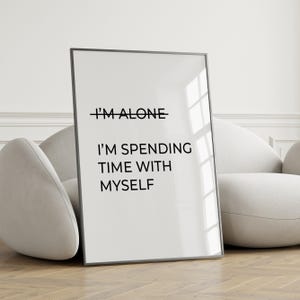 May include: A framed print with the text "I'M ALONE" crossed out, replaced with "I'M SPENDING TIME WITH MYSELF" in black font. The print is in a black frame and leans against a white sofa.