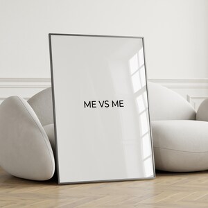 May include: A framed art print with the text "ME VS ME" in black, leaning against a white curved sofa. The frame is black, and the background is a light-colored wall and wooden floor.