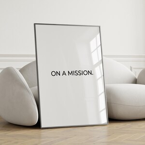 May include: A framed print with the text "ON A MISSION." The black text is centered on a white background. The frame is black and the print is leaning against a light-colored, curved sofa. The background is a light-colored wall and wooden floor.