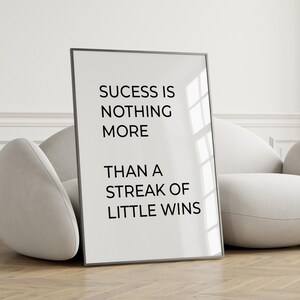 May include: A framed print with the text "Success is nothing more than a streak of little wins" in black font. The print is displayed against a neutral background, leaning against a white armchair.