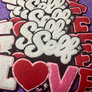 May include: A collection of chenille patches featuring the text "I LOVE SELF" and "LOVE" in various colors including white, red, and pink. The patches have a textured, fuzzy surface with black outlines and small star accents, layered on top of each other.