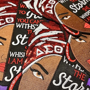 May include: Embroidered patches featuring a woman's face with brown skin, brown eyes, and red lips. The patches have a red and white sequined head covering and the Greek letters ΔΣΘ. Text includes "Whispered back I am the storm".