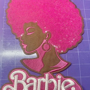 May include: An embroidered patch featuring a profile of a woman with a pink sequined afro and large hoop earrings. The word "Barbie" is embroidered in white script on a pink and brown background.