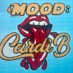 Może przedstawiać: Haftowane naszywki z napisami "MOOD: Cardi B" i grafiką czerwonych ust z wystającym językiem. Naszywki mają złotą i białą kolorystykę i znajdują się na niebieskiej powierzchni.