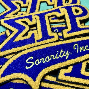 May include: Close-up of several blue and gold embroidered patches. The patches feature Greek letters and the words "Sorority, Inc." in a script font. The patches have a felt-like texture and a black border.
