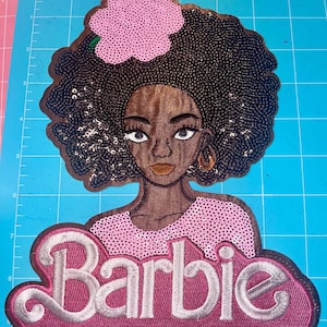 Puede incluir: Un parche con lentejuelas que presenta una ilustración estilizada de una mujer con piel oscura, que lleva una blusa rosa y una flor rosa en el pelo. La palabra "Barbie" está bordada en plata y rosa debajo de la imagen de la mujer.