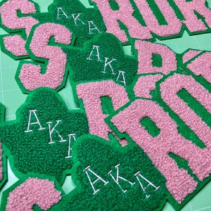 以下が含まれることがあります： ピンクとグリーンの刺繍パッチのコレクション。パッチは、文字「AKA」と「SOROR」をふわふわしたテクスチャスタイルで表示します。パッチは緑色の表面にあります。