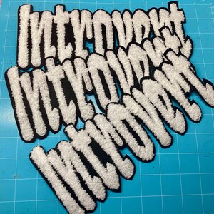 Könnte beinhalten: Eine Ansammlung von vier weißen Chenille-Aufnähern mit schwarzem Filzrand, die jeweils das Wort "introvert" in einer fetten, runden und blasenartigen Schrift zeigen. Die Aufnäher haben eine strukturierte, flauschige Oberfläche und sind überlappend angeordnet.