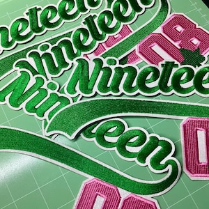 May include: Embroidered patches with the word "Nineteen" in green script with a white outline. Also, patches with the number "08" in pink with a white outline and a green leaf. Patches are on a green surface.