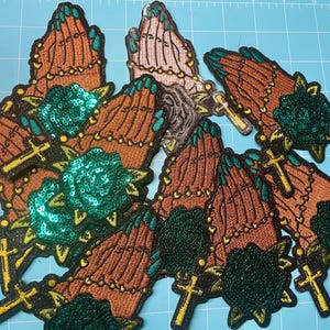 May include: Embroidered patches depicting praying hands with brown and teal details, gold crosses, and green sequined roses. The patches have a black border and are arranged on a blue cutting mat.