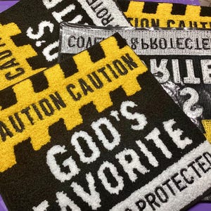 Könnte beinhalten: Mehrere schwarze und gelbe Matten mit Vorsichtsmotiven und weißem Text. Eine Matte zeigt "GOD'S FAVORITE PROTECTED". Andere zeigen "CAUTION". Die Matten haben eine strukturierte, plüschige Oberfläche.