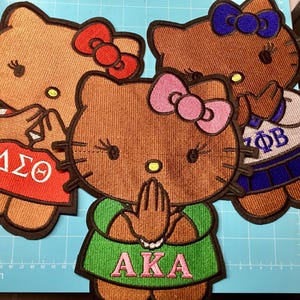 May include: Three embroidered patches featuring cartoon cat characters. One wears a red shirt with Greek letters ΔΣΘ and a red bow, one wears a blue and white outfit with Greek letters ΖΦΒ and a blue bow, and one wears a green shirt with AKA and a pink bow.
