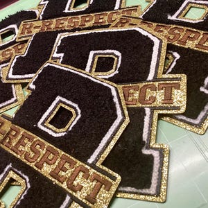 May include: Close-up of several black felt letter patches with a white border and a gold glitter accent. Each patch features the letter "R" and the word "RESPECT" in a gold glitter banner.