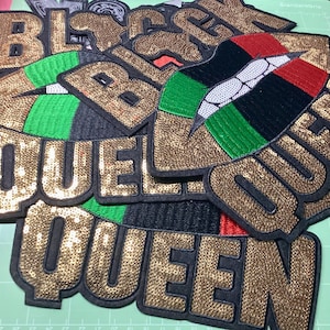 May include: A collection of sequined patches with the words "BLACK QUEEN" in gold sequins. Some patches feature a design with red, black, green, and white colors. The patches are on a green cutting mat.