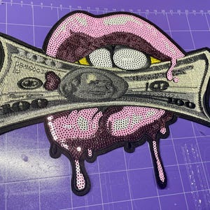 Op de afbeelding: Geborduurde patch met een mond met roze lippen en een druipend effect, met een gedetailleerde $100-biljet. De patch heeft een zwarte rand en is versierd met pailletten.