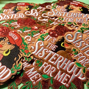 May include: Pile of embroidered patches with the text "It's the Sisterhood for Me!" and an illustration of a woman wearing a red headscarf. The patches are brown, red, green, and white.