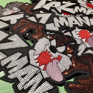 May include: A collection of embroidered patches featuring the Tasmanian Devil cartoon character. The patches have a black background with white sequin-covered letters spelling "TAZ MANIA". The Tasmanian Devil is depicted with a red mouth and a purple tongue.