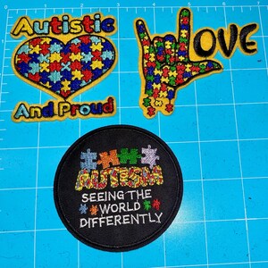 Puede incluir: Tres parches bordados con temas relacionados con el autismo. Uno es un parche en forma de corazón con las palabras "Autistic And Proud". Otro parche muestra una mano haciendo el signo "Te amo". El tercer parche es redondo y dice "Autism Seeing The World Differently".