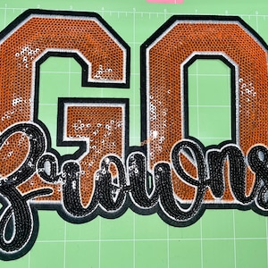 Pode incluir: Um grande patch bordado com as palavras "GO Browns" em lantejoulas laranja e pretas. As letras "GO" estão em um estilo de bloco, enquanto "Browns" está em uma fonte cursiva. O patch tem uma borda preta e um contorno branco.