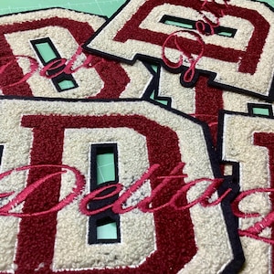 Może przedstawiać: Zbliżenie kilku naszywek z literami uniwersyteckimi. Każda naszywka ma dużą, bordową literę „D” z kremowym tłem i czarnym obramowaniem. Słowo „Delta” jest wyhaftowane kursywą.