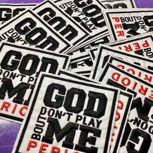 以下が含まれることがあります： 白地に黒と赤の文字が刺繍されたパッチの山。「GOD DON'T PLAY ABOUT ME PERIOD」と書かれています。パッチは黒い縁取りがあり、紫色の表面に積み重ねられています。
