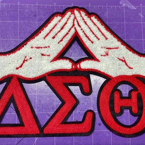 May include: A chenille patch featuring two white hands forming a triangle shape above the Greek letters Delta, Sigma, and Theta in red. The patch has a black border and is positioned on a purple cutting mat with visible inch markings.