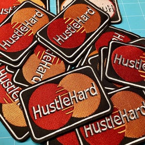 May include: A collection of rectangular embroidered patches featuring a black background with two overlapping circles in red and orange. The text HustleHard is embroidered in white across the center of each patch, mimicking a credit card logo design.