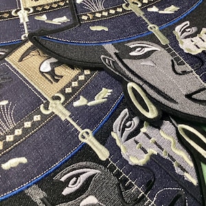 May include: Embroidered patches with an Egyptian pharaoh design. The patches have a denim-like background with detailed embroidery in grey, beige, and blue. The design includes hieroglyphs and stylized figures.
