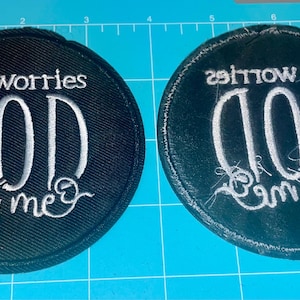 May include: Two round, black embroidered patches with white text. One patch reads "no worries GOD got me" in a script font. The other patch has the same text, reversed. Patches are on a blue surface.