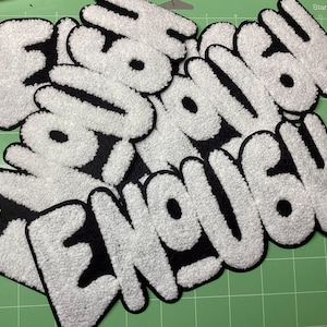 May include: A collection of white, fluffy, embroidered patches with the word "ENOUGH" in a bold, bubble-style font. The patches have a black border and are arranged on a green cutting mat.