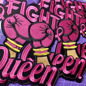 Peut inclure: Plusieurs écussons brodés représentant une paire de gants de boxe roses avec des bandes de poignet jaunes et un ruban de sensibilisation rose. Le texte FIGHT est brodé en rose au-dessus des gants et le mot QUEEN est brodé en rose en dessous.