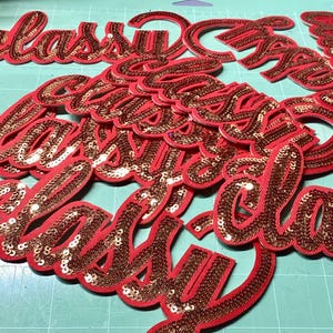 Może przedstawiać: Czerwone i złote naszywki "classy" z cekinami. Naszywki są w kursywnym kroju pisma z czerwoną obwódką i wypełnione złotymi cekinami. Naszywki są rozrzucone na zielonej macie do cięcia.