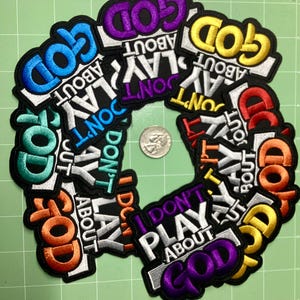 May include: A selection of embroidered patches, each bearing the phrase "I DON'T PLAY ABOUT GOD" in diverse colours. The patches are black with white and vibrant text, including blue, purple, yellow, and orange. A coin is in the centre.
