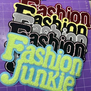 May include: A collection of embroidered patches with the word "Fashion" in various colors, including black, gray, red, and yellow. One patch reads "Fashion Junkie" in green and blue. The patches have a fuzzy, textured appearance.