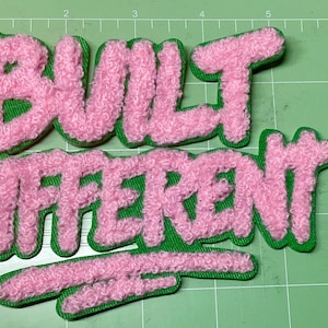 May include: Pink, fluffy patches spelling "BUILT DIFFERENT" in a bubble letter style, with green edging. A coin is next to the patches for scale. The patches are on a green cutting mat.