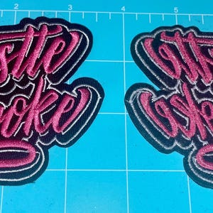 May include: Two embroidered patches with the phrases "Hustle or Be Broke" and "It's Built, Not Bought" in a cursive font. The patches have a black background with pink and white lettering.