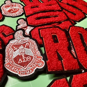 May include: A collection of red and black embroidered patches and letters. The patches feature a crest design with white and red detailing. The letters are in a bold, block style. The items are scattered on a green surface.