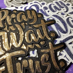 May include: Embroidered patches with the words "Pray, Wait, Trust" in gold sequins on a black background. Other patches feature the same words in white with blue sequin accents. The patches are ideal for clothing or accessories.