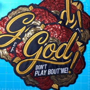 May include: A large embroidered patch featuring a cluster of red and gold sequined roses. The word God is stitched in large gold cursive letters, with the text DON'T PLAY BOUT'ME! in white block letters underneath on a black fabric background.