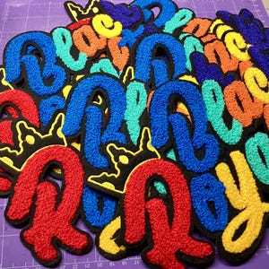 May include: A collection of colorful, embroidered patches spelling out the word "Rebel" in a playful, bubble-letter style. The patches feature a black border and are in shades of red, blue, yellow, and orange.