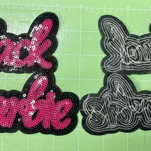 Peut inclure: Deux écussons avec le texte "Black Barbie". L'un est rose avec des paillettes et un bord noir. L'autre est noir avec des coutures blanches. Les deux sont sur une surface verte.