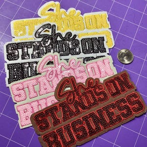 Puede incluir: Primer plano de varios parches bordados con la frase "She Stands On Business". Los parches son de varios colores, incluyendo dorado, negro, rosa y rojo, con detalles de lentejuelas.