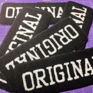 May include: A collection of black, rectangular patches, each bearing the word "ORIGINAL" in white, textured lettering. The patches are overlapping, with the text prominent. The background is a purple cutting mat.