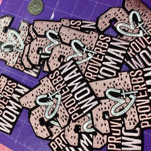 Könnte beinhalten: Eine Sammlung gestickter Aufnäher mit dem Schriftzug "PROVERBS WOMAN" in Weiß auf schwarzem Hintergrund. Jeder Aufnäher hat einen rosa Hintergrund mit einem Herz- und Kreuz-Design. Eine Münze ist sichtbar.