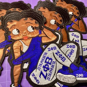 May include: Embroidered patches featuring a cartoon woman in a blue dress and white pants with the text "ZETA PHI BETA". The patches have a black border and are on a purple surface. The woman has black hair and blue earrings.