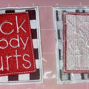 Puede incluir: Dos parches cuadrados de tela. Uno tiene un cuadrado rojo con las palabras "BACK & BODY HURTS" bordadas en blanco. El otro tiene el mismo diseño, pero el texto es blanco sobre fondo blanco. Ambos tienen un borde a cuadros blanco y negro.