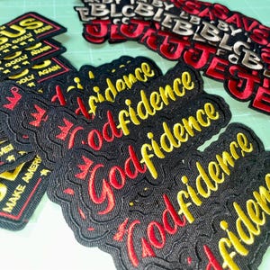 May include: Embroidered patches with phrases in gold and red thread on a black background. Phrases include "JESUS", "MAKE AMERICA GODLY AGAIN", and "Godfidence". The patches are rectangular with a decorative border.