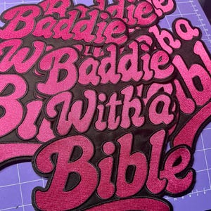 May include: Several embroidered patches with the phrase "Baddie With a Bible" in a retro font. The patches feature a black background with pink lettering and a pink border. The patches are stacked on top of each other.