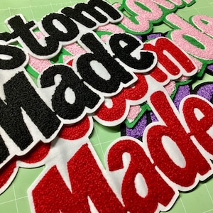 May include: Pile of custom-made embroidered patches in various colors, including red, black, pink, and purple. The patches feature the words "custom" and "made" in a bold, textured font with white borders. The patches are arranged on a green surface.