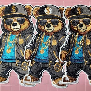 May include: Three identical embroidered patches featuring a brown bear wearing a black hoodie, black pants, and a black baseball cap with a gold dollar sign. The bear wears black sunglasses and gold chain necklaces over a bright blue shirt.